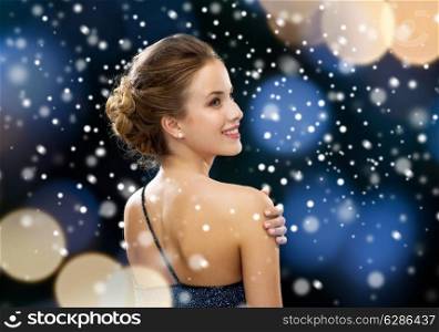 people, christmas, holidays and glamour concept - smiling woman in evening dress over black background over night lights and snow background from back