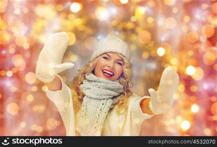people, christmas, holidays and new year concept - happy smiling beautiful woman in winter hat, scarf and mittens showing her palms over holidays lights and snow background