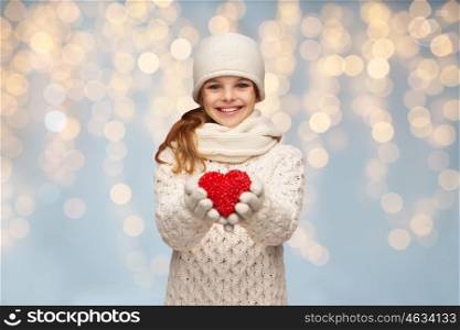 people, christmas, holidays, charity and love concept - smiling teenage girl in winter clothes with small red heart over lights background