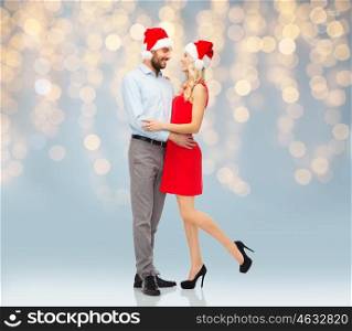 people, christmas, love, couple and holidays concept - happy young woman and man in santa hats hugging over lights background