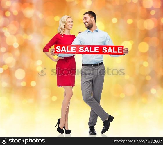 people, christmas, sale, discount and holidays concept - happy couple with red sale sign looking at each other over lights background