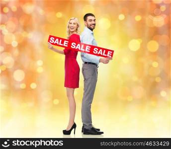 people, christmas, sale, discount and holidays concept - happy couple with red sale sign standing back to back over lights background
