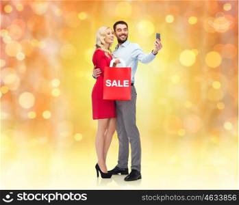 people, christmas, sale, technology and holidays concept - happy young woman and man with red shopping bag taking selfie by smartphone over lights background