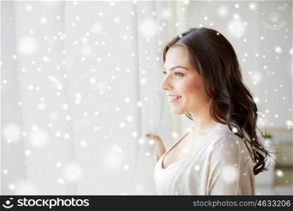 people, christmas, winter and leisure concept - happy young woman looking through window at home over snow