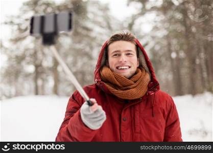 people, christmas, winter and season concept - happy smiling man in hood and scarf taking picture by smartphone selfie stick outdoors