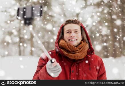 people, christmas, winter and season concept - happy smiling man in hood and scarf taking picture by smartphone selfie stick outdoors. happy man taking selfie by smartphone in winter