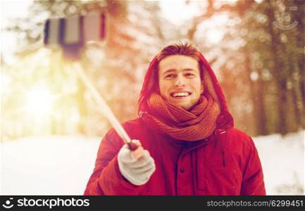 people, christmas, winter and season concept - happy smiling man in hood and scarf taking picture by smartphone selfie stick outdoors. happy man taking selfie by smartphone in winter