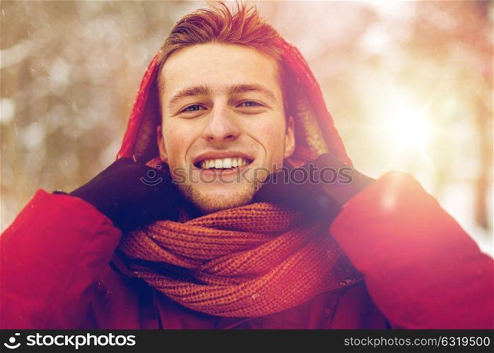 people, christmas, winter and season concept - happy smiling man in jacket with hood adjusting scarf outdoors. happy man in winter jacket and scarf outdoors