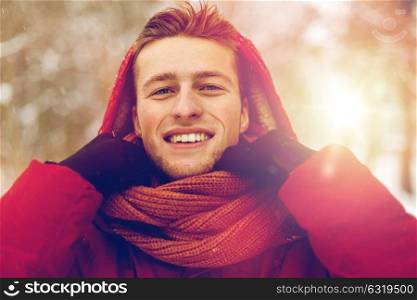 people, christmas, winter and season concept - happy smiling man in jacket with hood adjusting scarf outdoors. happy man in winter jacket and scarf outdoors