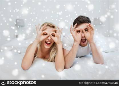 people, christmas, winter holidays, love and fun concept - happy couple lying in bed at home and making finger glasses over snow