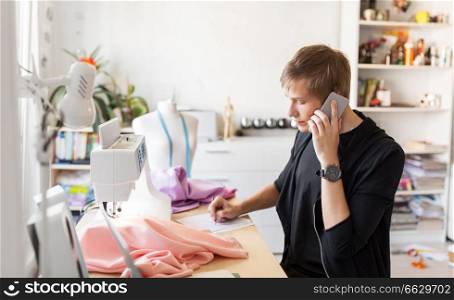 people, clothing and tailoring concept - fashion designer calling on smartphone and making notes at studio. fashion designer calling on smartphone at studio
