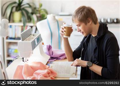 people, clothing and tailoring concept - fashion designer drinking coffee from disposable paper cup and reading book at studio. fashion designer with coffee and book at studio