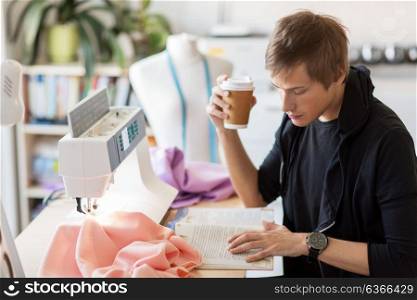 people, clothing and tailoring concept - fashion designer drinking coffee from disposable paper cup and reading book at studio. fashion designer with coffee and book at studio
