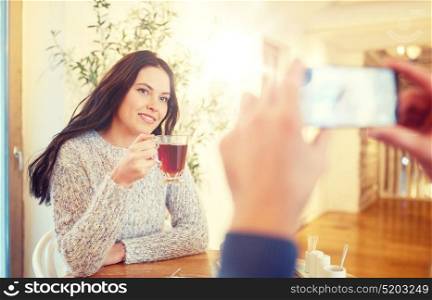 people, communication and dating concept - man with smartphone taking picture of woman drinking tea at cafe or restaurant. man taking picture of woman by smartphone at cafe