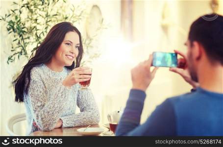 people, communication and dating concept - man with smartphone taking picture of woman drinking tea at cafe or restaurant. man taking picture of woman by smartphone at cafe