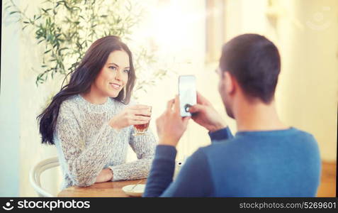 people, communication and dating concept - man with smartphone taking picture of woman drinking tea at cafe or restaurant. man taking picture of woman by smartphone at cafe