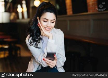People, communication and lifestyle concept. Happy brunette woman with attractive face looking elegantly making video call, using online app on her mobile phone or listening to music relaxing indoors