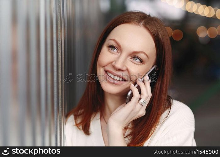 People, communication and technology concept. Close up shot of dreamy young European woman with staright dyed hair, keeps smart phone near ear, being in good mood, has make up, positive smile