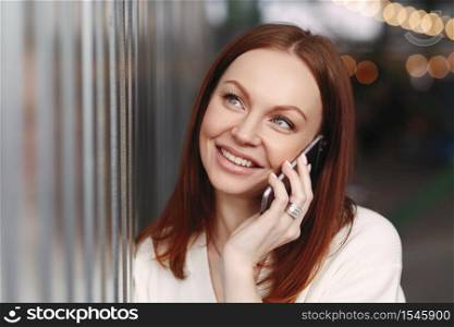 People, communication and technology concept. Close up shot of dreamy young European woman with staright dyed hair, keeps smart phone near ear, being in good mood, has make up, positive smile