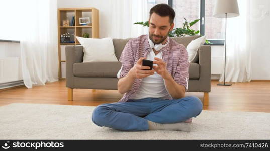 people, communication, technology and internet concept - smiling man with headphones on his neck using smartphone at home. smiling man using smartphone at home