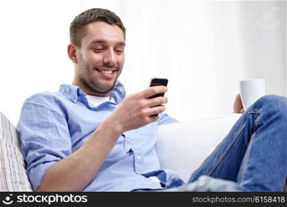 people, communication, technology and internet concept - smiling man with smartphone texting or reading message and drinking tea at home