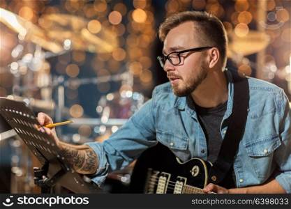 people, composition and entertainment concept - man with guitar writing lyrics or notes to music book at studio over holidays lights background. man with guitar writing to music book at studio