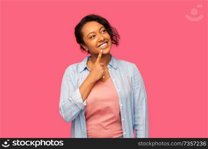 people concept - happy african american young woman pointing finger to her lips or thinking over pink background. african american woman looking un up and thinking