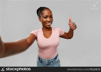 people concept - happy african american young woman taking selfie and showing thumbs up over grey background. african woman taking selfie and showing thumbs up