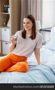 people concept - happy smiling girl with glass of water sitting on bed at home. girl with glass of water sitting on bed at home