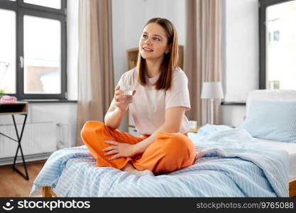 people concept - happy smiling girl with glass of water sitting on bed at home. girl with glass of water sitting on bed at home