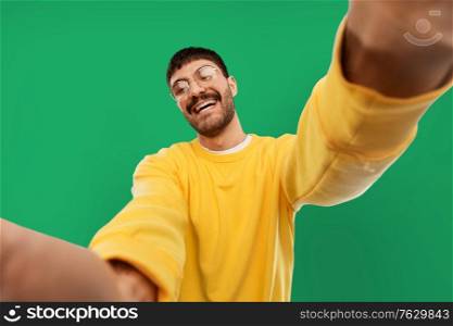 people concept - happy smiling young man in glasses and yellow sweatshirt making selfie over emerald green background. happy young man in yellow sweatshirt making selfie