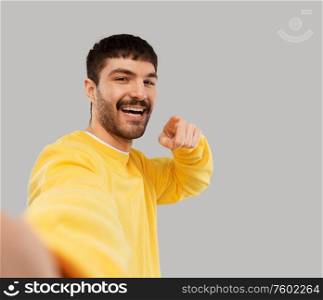 people concept - happy smiling young man in yellow sweatshirt making selfie and pointing finger to camera over grey background. happy young man in yellow sweatshirt making selfie