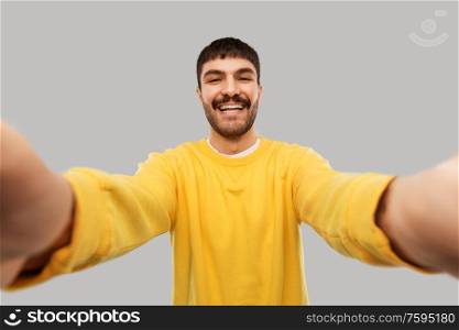 people concept - happy smiling young man in yellow sweatshirt making selfie over grey background. happy young man in yellow sweatshirt making selfie