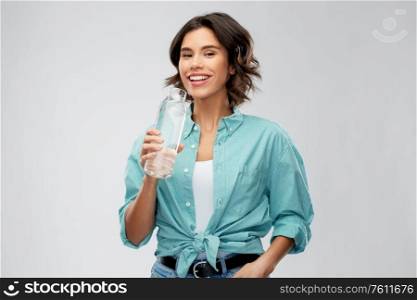 people concept - portrait of happy smiling young woman in turquoise shirt drinking water from reusable glass bottle grey background. smiling woman drinking water from glass bottle