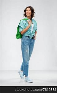 people concept - portrait of happy smiling young woman in turquoise shirt and jeans walking with green reusable canvas bag for food shopping on grey background. woman with reusable canvas bag for food shopping