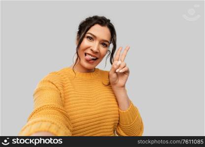people concept - portrait of happy smiling young woman with pierced nose taking selfie showing peace gesture and tongue over grey background. woman with pierced nose taking selfie shows peace