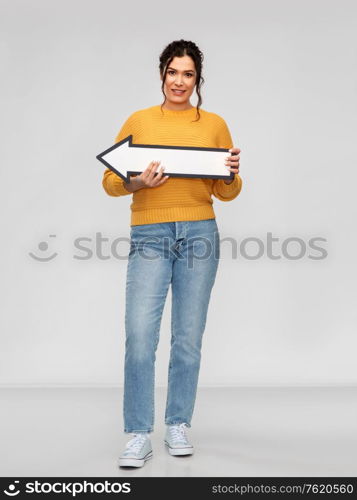 people concept - portrait of happy smiling young woman with pierced nose holding big white thick arrow showing to left over grey background. happy young woman with big white thick left arrow