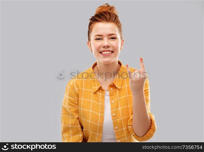 people concept - smiling red haired teenage girl in checkered shirt showing rock gesture over grey background. happy red haired teenage girl showing rock gesture