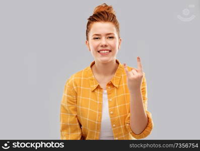 people concept - smiling red haired teenage girl in checkered shirt showing rock gesture over grey background. happy red haired teenage girl showing rock gesture