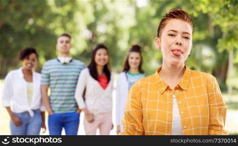 people concept - smiling red haired teenage girl in checkered shirt showing her tongue over group of friends at summer park background. smiling red haired teenage girl showing her tongue