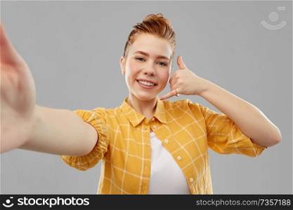 people concept - smiling red haired teenage girl in checkered shirt taking selfie and making calling gesture by hand over grey background. teenage girl taking selfie making calling gesture