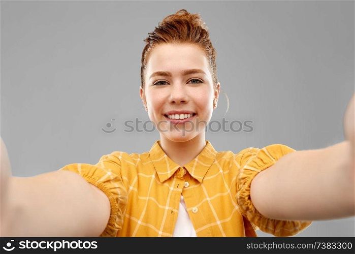 people concept - smiling red haired teenage girl in checkered shirt taking selfie over grey background. smiling red haired teenage girl taking selfie