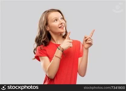 people concept - smiling teenage girl with long hair in red t-shirt pointing fingers to something over grey background. smiling teenage girl pointing fingers to something