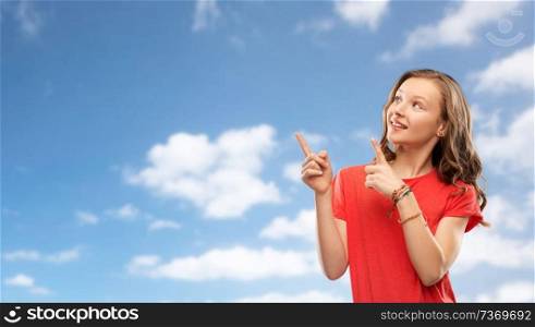 people concept - smiling teenage girl with long hair in red t-shirt pointing fingers to something over blue sky and clouds background. smiling teenage girl pointing fingers to something