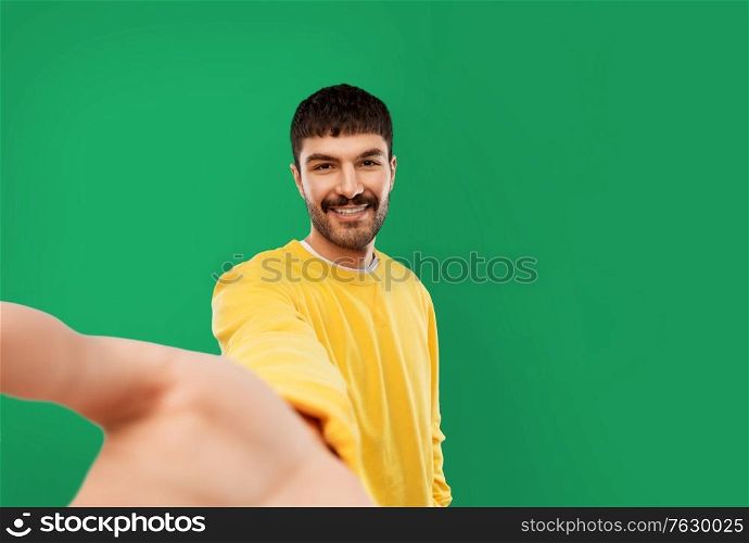 people concept - smiling young man in yellow sweatshirt making selfie over emerald green background. happy young man in yellow sweatshirt making selfie