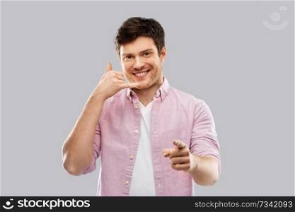 people concept - smiling young man showing phone call gesture and pointing to you over grey background. smiling young man showing phone call gesture