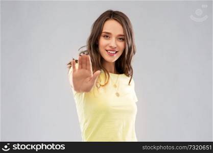 people concept - smiling young woman or teenage girl in blank yellow t-shirt showing stop gesture over grey background. young woman or teenage girl showing stop gesture