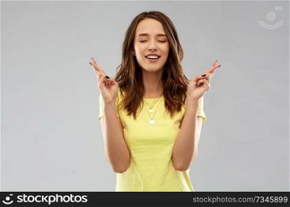 people concept - smiling young woman or teenage girl in blank yellow t-shirt holding fingers crossed over grey background. young woman or teenage girl with fingers crossed