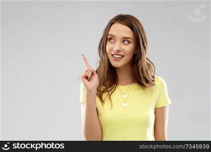 people concept - smiling young woman or teenage girl in blank yellow t-shirt pointing finger up over grey background. young woman or teenage girl pointing finger up