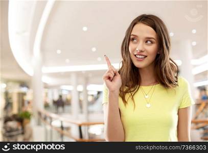 people concept - smiling young woman or teenage girl in blank yellow t-shirt pointing finger up over shopping mall background. teenage girl pointing finger up in shopping mall